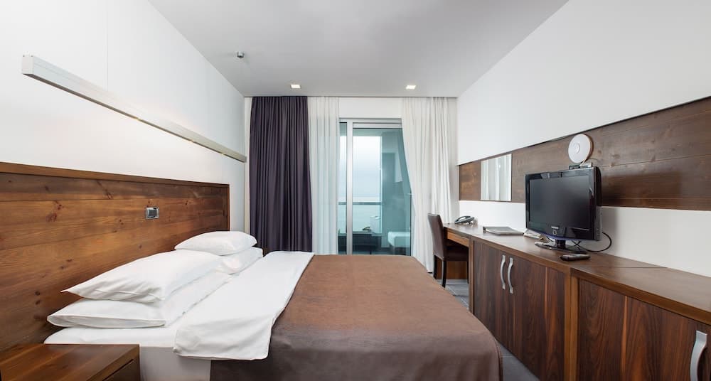 A cozy hotel room with a neatly made bed, wooden accents, a desk with a TV, and a balcony offering a serene view, ideal for travelers seeking comfort and relaxation.