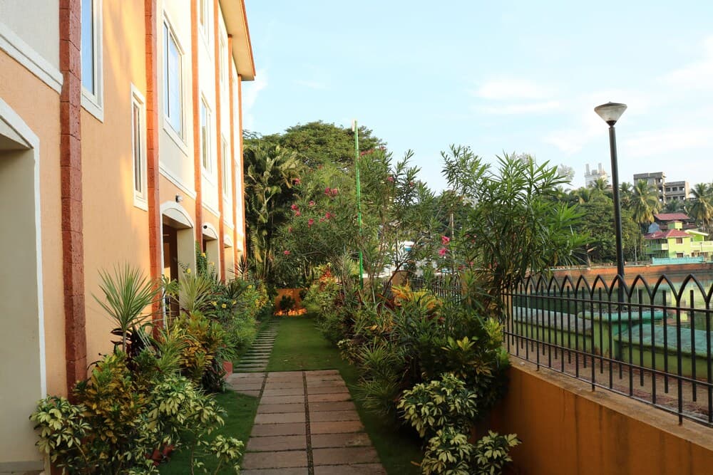 A serene residential pathway lined with lush greenery and vibrant plants leads to a tranquil waterfront view, ideal for travelers seeking peaceful accommodations in a tropical setting.
