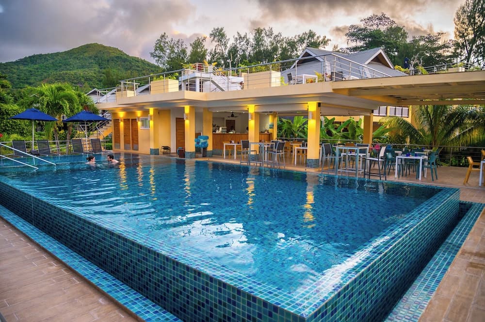 A serene tropical resort features a vibrant blue-tiled pool surrounded by lounge chairs under umbrellas, with a warmly lit dining area and lush greenery in the background, ideal for a relaxing vacation getaway.