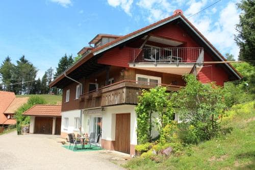 A charming red-roofed house nestled on a lush hillside under a bright blue sky, featuring balconies and outdoor seating, ideal for a serene countryside getaway.