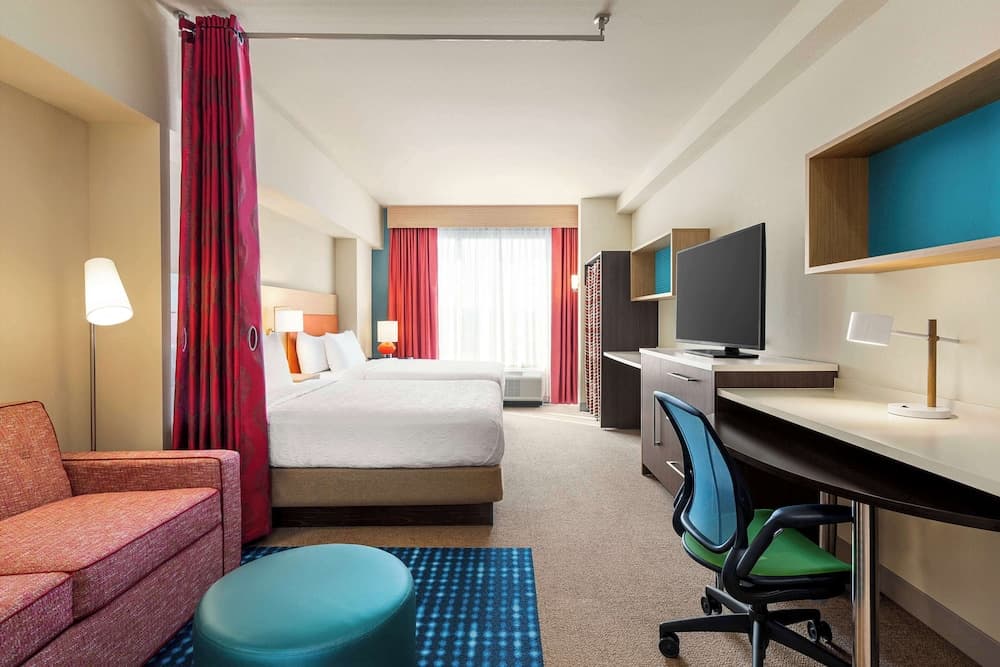 A cozy hotel room features vibrant red curtains, a comfortable bed, a modern desk setup with a blue chair, and a plush sofa, ideal for travelers seeking a blend of relaxation and productivity.