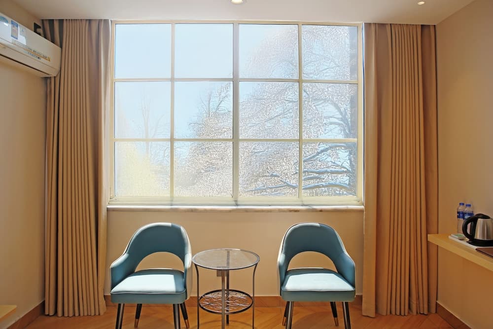 A cozy hotel room corner features two blue chairs flanking a glass table by a large frosted window overlooking leafless trees, ideal for travelers seeking a serene and inviting space for relaxation or work.