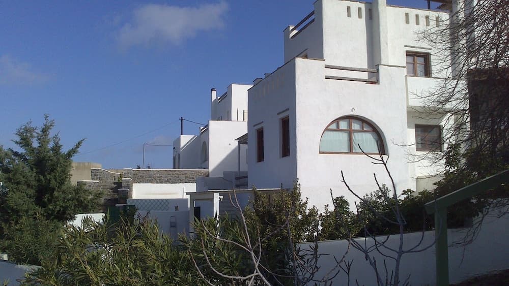 A serene Mediterranean village scene features white-washed buildings with arched windows under a clear blue sky, surrounded by lush greenery, ideal for travelers seeking a peaceful coastal retreat.