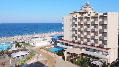 A vibrant seaside resort features a modern hotel complex with balconies overlooking the calm blue sea, surrounded by lush greenery and outdoor amenities, ideal for a relaxing beach vacation.