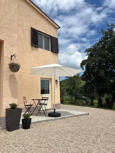 A serene countryside retreat features a cozy patio with a white umbrella, chairs, and potted plants against a rustic building under a partly cloudy sky, ideal for travelers seeking relaxation in a tranquil rural setting.