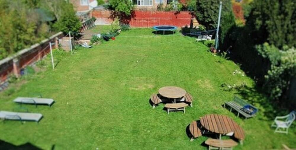 A serene suburban backyard features lush green grass, wooden picnic tables, lounge chairs, and a trampoline, ideal for family gatherings or relaxing outdoor activities.