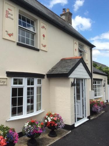 A charming, pastel-colored cottage adorned with vibrant flower pots and quaint window decorations invites travelers to explore its cozy ambiance in a picturesque village setting.