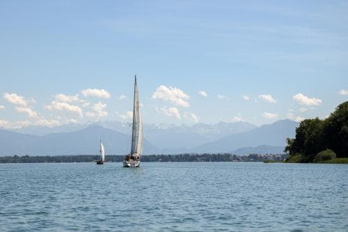 A serene lakeside scene features sailboats gliding on calm waters under a partly cloudy sky, with distant mountains enhancing the tranquil atmosphere, ideal for leisurely sailing trips or nature photography.
