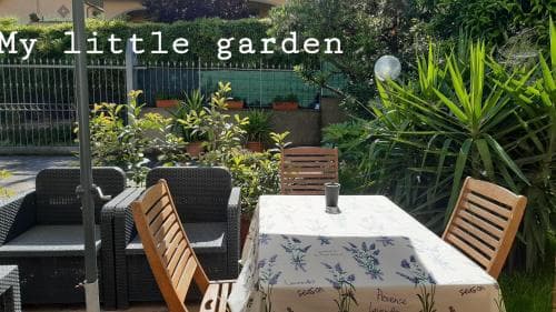 A cozy backyard garden features a table set with a floral tablecloth surrounded by wooden chairs, lush greenery, and potted plants, creating a serene and inviting atmosphere perfect for relaxing or hosting small gatherings.