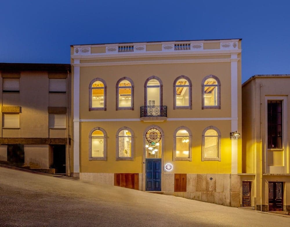 A warmly lit, historic yellow building with arched windows and a blue door stands invitingly on a quiet street at dusk, perfect for travelers seeking charming accommodations in a serene urban setting.