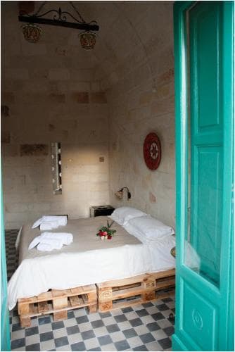 A cozy bedroom with a rustic pallet bed, checkered floor, and turquoise door evokes a serene, eco-friendly retreat ideal for travelers seeking unique accommodations in a charming, historic setting.