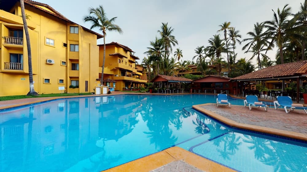 A serene tropical resort features a vibrant blue pool surrounded by palm trees, yellow buildings, and lounge chairs, ideal for vacation relaxation and leisure photography.