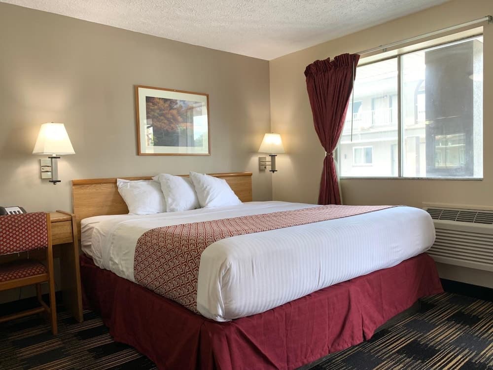A cozy hotel room features a neatly made bed with red accents, framed artwork, wall-mounted lamps, and a window with burgundy curtains, ideal for travelers seeking comfort and relaxation.