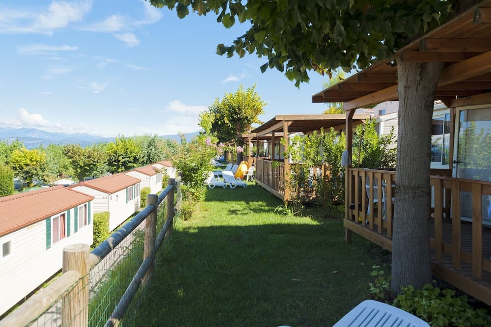 A serene campground with cozy cabins, lush greenery, wooden pergolas, and lounge chairs under a clear blue sky, perfect for a tranquil family vacation.