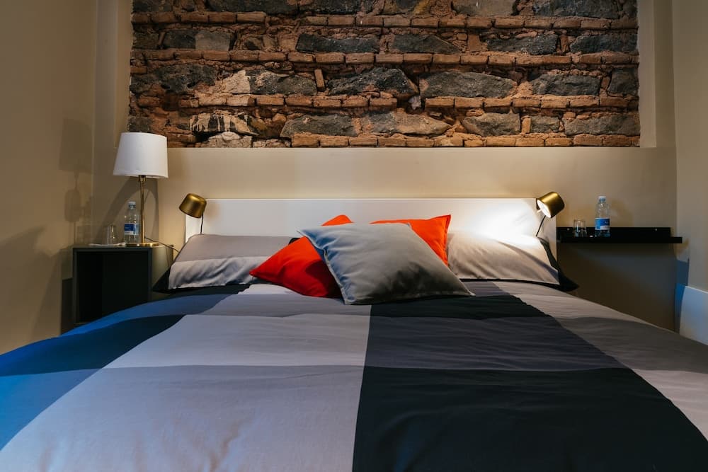 A cozy hotel room features a neatly made bed with colorful pillows against a rustic stone wall backdrop, ideal for travelers seeking comfort and charm.