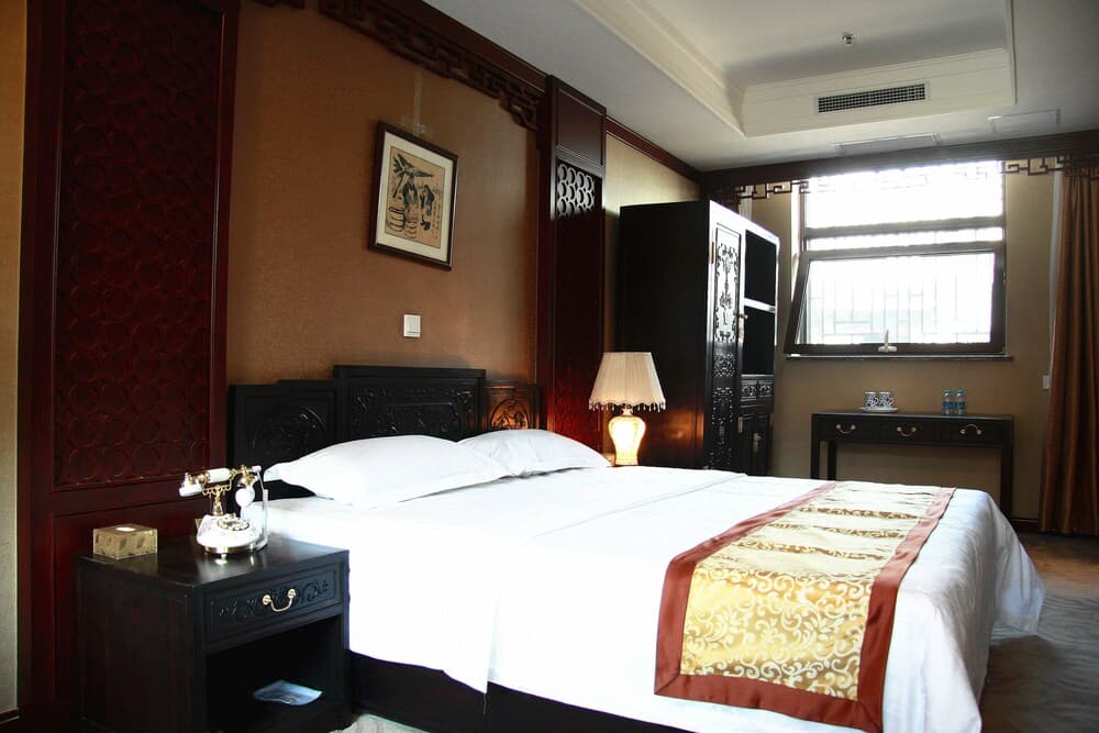 A cozy hotel room with traditional decor features a neatly made bed adorned with a golden runner, vintage bedside telephone, ornate wooden furnishings, and soft lighting, ideal for travelers seeking comfort and cultural ambiance.
