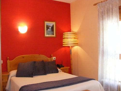 A cozy hotel room features a neatly made bed against a vibrant red accent wall, complemented by soft lighting from a striped floor lamp and a warm sconce, ideal for travelers seeking comfort and relaxation.