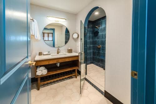 A cozy hotel bathroom features hexagonal tiled floors, a wooden vanity with neatly folded towels, a round mirror, and a striking blue-tiled shower under an arched doorway, ideal for travelers seeking comfort and style.