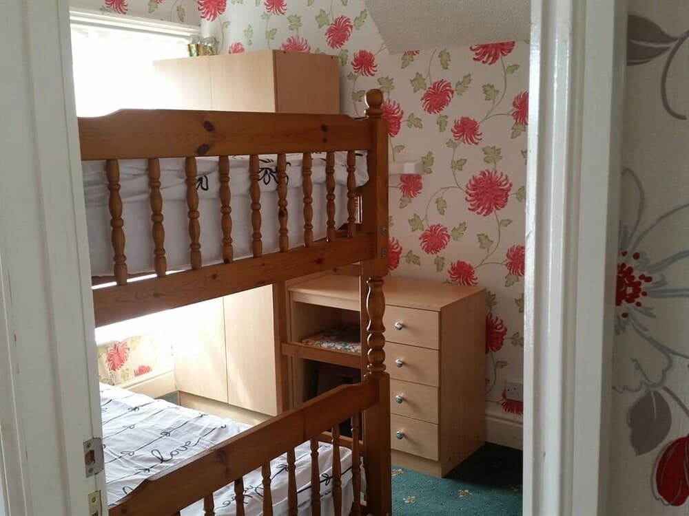 A cozy children's bedroom features a wooden bunk bed with floral wallpaper, a chest of drawers, and natural light streaming through the window, ideal for family vacation accommodations.