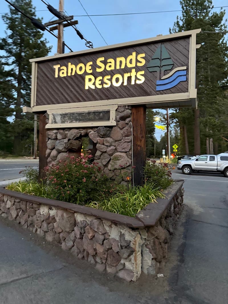 A welcoming entrance sign for Tahoe Sands Resort stands amidst lush greenery and towering pines, inviting travelers to explore its serene lakeside retreat in Tahoe Vista, California.