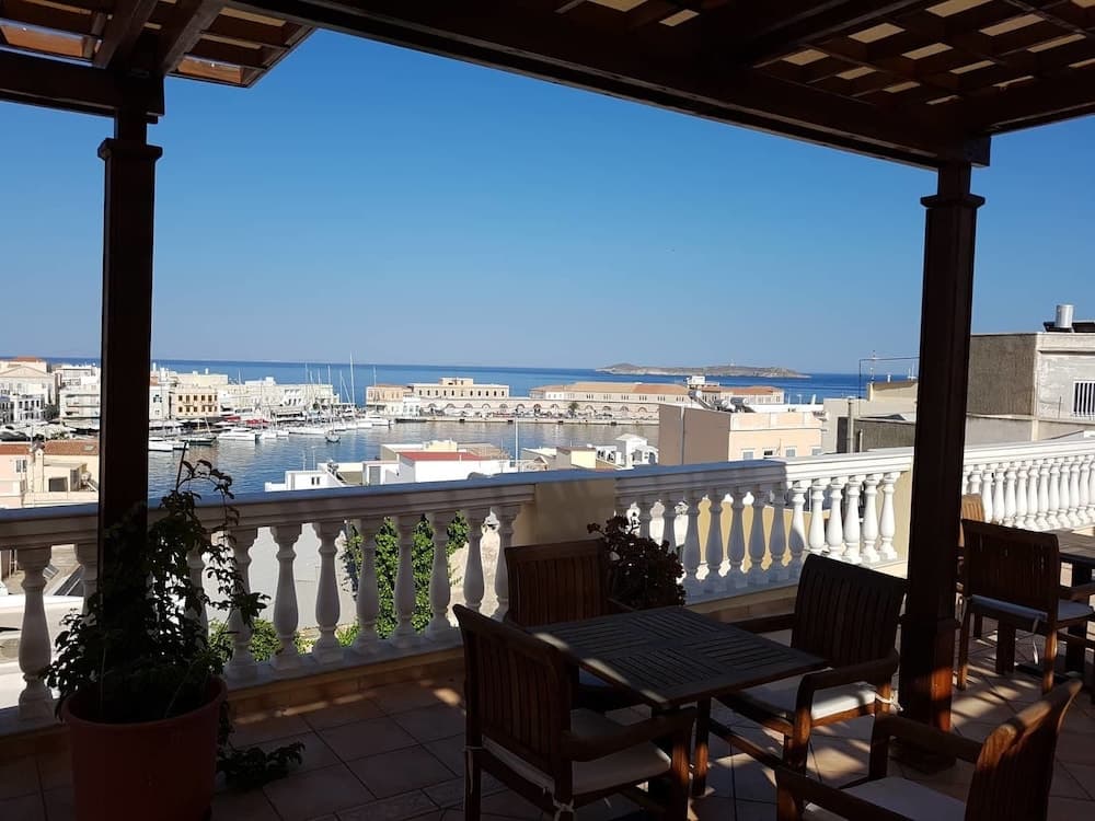 A serene balcony with wooden furniture overlooks a picturesque harbor filled with boats and surrounded by white buildings under a clear blue sky, ideal for a relaxing seaside getaway.