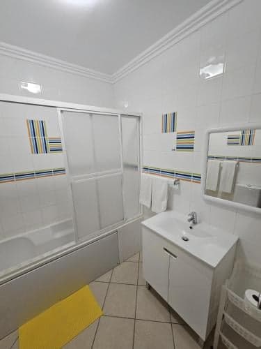A clean, modern bathroom with striped tile accents, a glass-enclosed bathtub, white vanity, and yellow bath mat creates a bright and inviting atmosphere ideal for travelers seeking comfort and functionality in their accommodations.