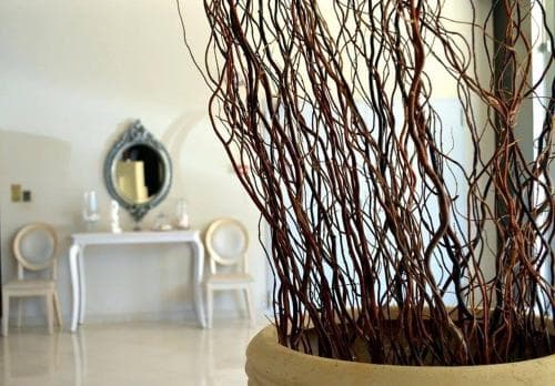 A serene indoor setting features a large pot of twisted branches in the foreground, with a white table adorned by candles and a decorative mirror, evoking a tranquil and elegant atmosphere ideal for a relaxing retreat or interior design inspiration.