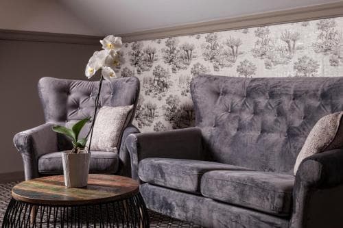 A cozy hotel lounge features plush gray velvet seating, a rustic coffee table with an orchid plant, and elegant tree-patterned wallpaper, creating a serene atmosphere ideal for relaxation during travel.