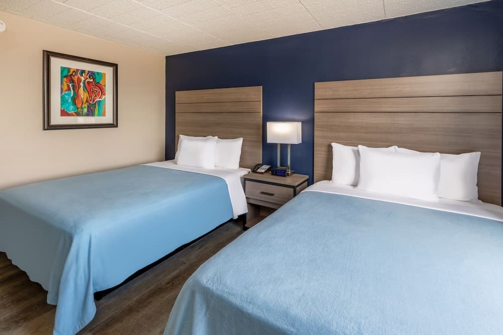 A cozy hotel room features two neatly made beds with blue linens, a wooden nightstand with a lamp and clock, a vibrant wall art piece, and a calming color scheme ideal for travelers seeking comfort and relaxation.
