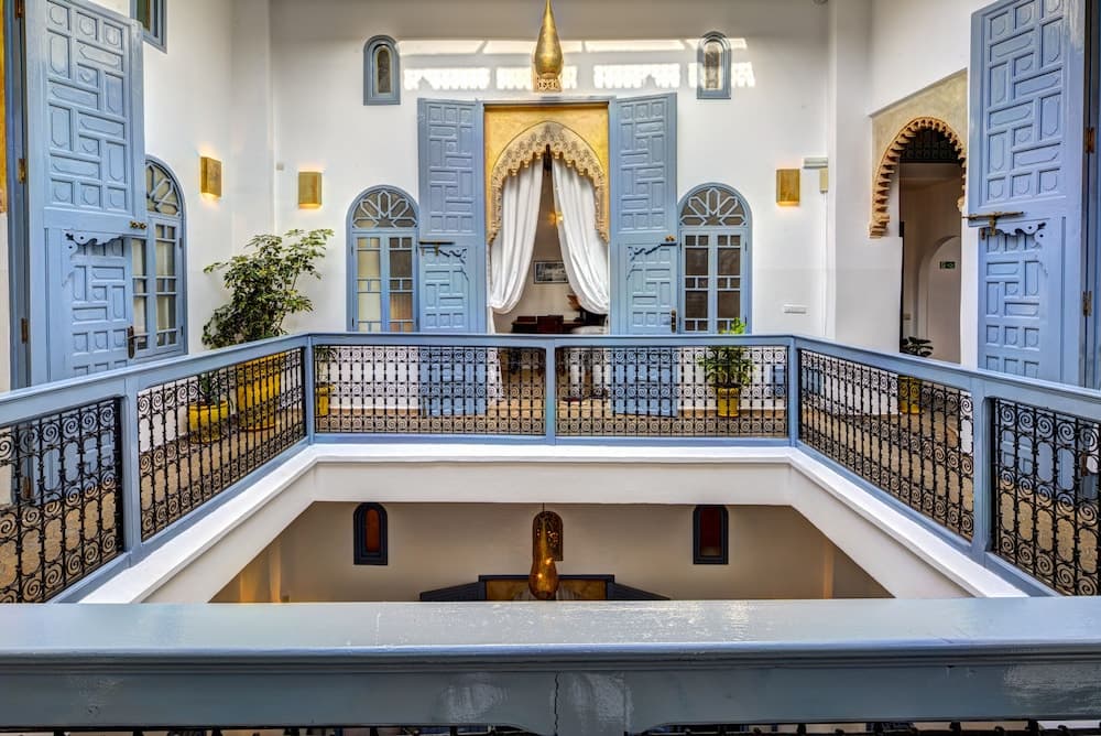 A serene courtyard in a traditional riad features ornate blue doors, intricate iron railings, and lush greenery, ideal for travelers seeking cultural immersion and relaxation.