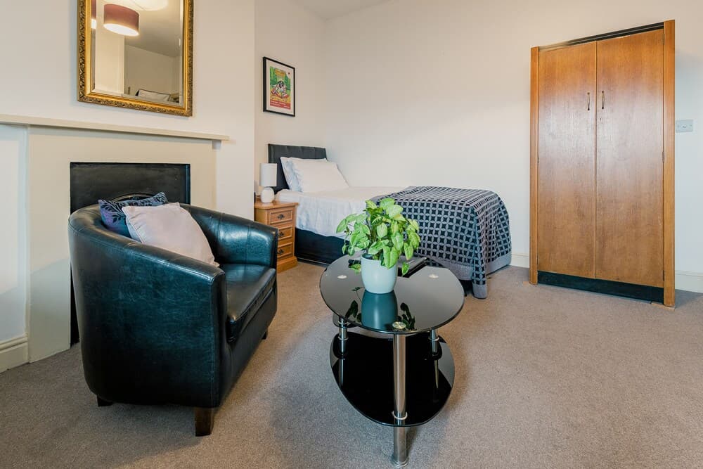 A cozy studio apartment features a black leather armchair, a glass coffee table with a green plant, a neatly made bed, and wooden furniture, creating a comfortable and inviting atmosphere ideal for short-term travelers seeking a homey stay.