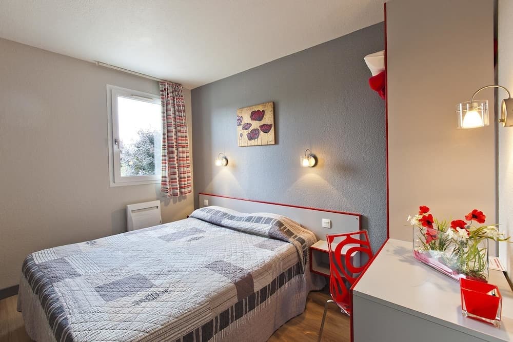 A cozy hotel room features a neatly made bed by a window with striped curtains, a gray wall adorned with floral art, and a vibrant red chair beside a desk with fresh flowers, ideal for travelers seeking comfort and style.