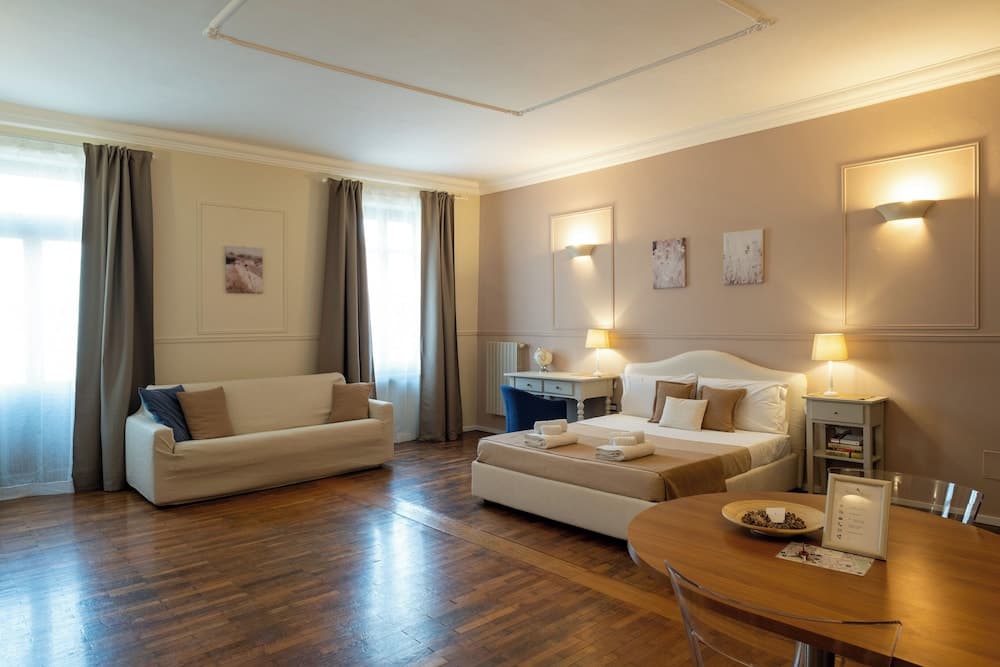A cozy hotel suite features warm wooden floors, a plush sofa by large windows, a neatly made bed with soft lighting, and a dining area, ideal for travelers seeking comfort and relaxation.
