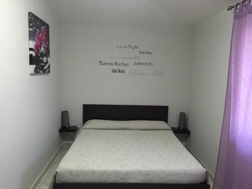 A cozy bedroom with a neatly made bed, purple curtains, and wall art featuring "Good Night" in multiple languages creates a serene atmosphere ideal for travelers seeking restful accommodations.