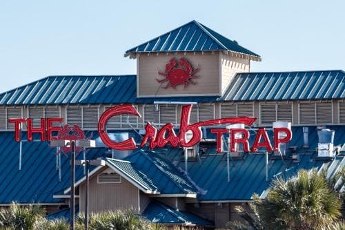 A vibrant seafood restaurant with a bold "The Crab Trap" sign stands amidst palm trees under a clear sky, inviting travelers to enjoy coastal cuisine in a relaxed beachside setting.