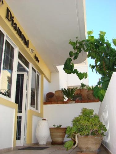 A sunny balcony adorned with lush potted plants and terracotta pots overlooks a charming yellow building entrance labeled "Diamond," ideal for travelers seeking a cozy and vibrant accommodation spot.