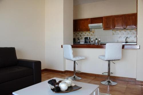 A cozy apartment interior features a modern kitchenette with wooden cabinets, white bar stools at the counter, and a comfortable seating area, ideal for travelers seeking a homey and functional stay.