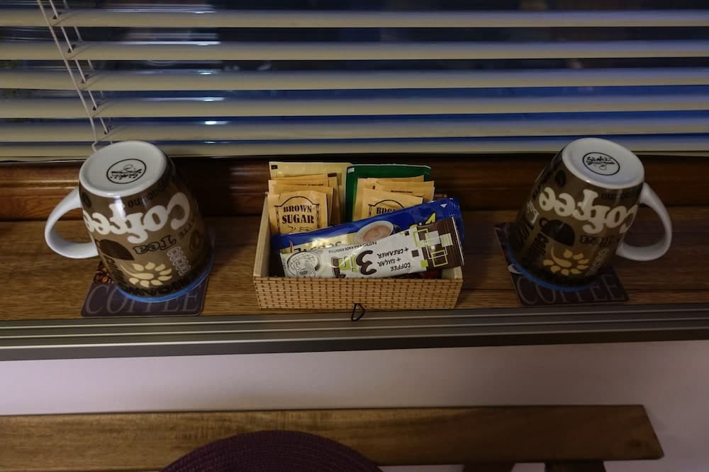 A cozy café corner features two coffee-themed mugs flanking a basket of sugar packets on a wooden ledge beneath blinds, ideal for travelers seeking a quiet spot to enjoy their morning brew.