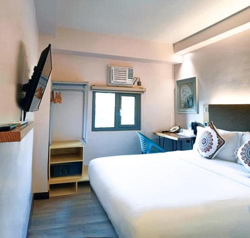A cozy hotel room with a neatly made bed, vibrant pillows, and a small desk by the window, exudes a welcoming ambiance ideal for travelers seeking comfort and convenience.