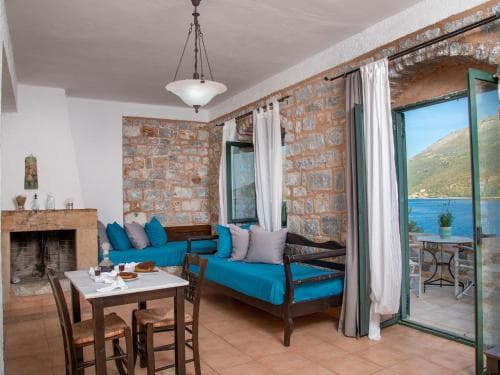 A cozy living room with stone walls, a fireplace, and blue sofas opens to a serene balcony overlooking the sea, ideal for a tranquil seaside retreat.