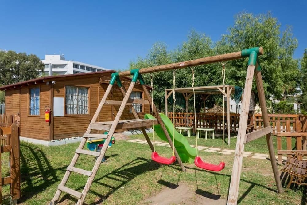 A vibrant playground with swings and a slide sits invitingly in front of a cozy wooden cabin under a clear blue sky, perfect for family-friendly vacation spots.