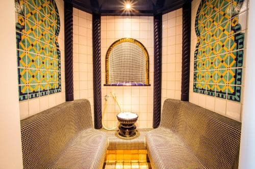 A cozy spa room features ornate tiled walls, plush seating, and a central fountain under warm lighting, ideal for relaxation and wellness retreats.