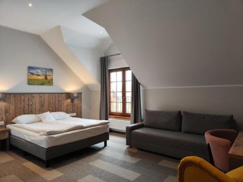 A cozy attic bedroom with a slanted ceiling features a neatly made bed, a comfortable sofa, and a bright window, creating an inviting atmosphere ideal for travelers seeking a peaceful retreat.