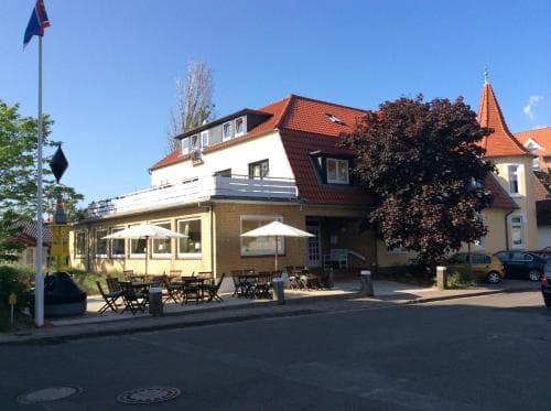 A quaint European café with outdoor seating under white umbrellas invites travelers to relax in the sunny town square, surrounded by charming buildings and a vibrant tree.