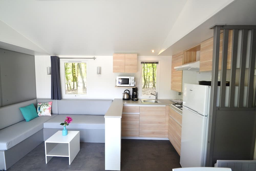 A cozy cabin interior features a compact kitchen with wooden cabinets, a small dining area with colorful cushions, and windows offering a serene forest view, ideal for a tranquil getaway.
