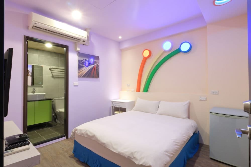 A cozy hotel room features a neatly made bed with colorful wall art, a modern bathroom with green accents, and ambient lighting, ideal for travelers seeking comfort and style.