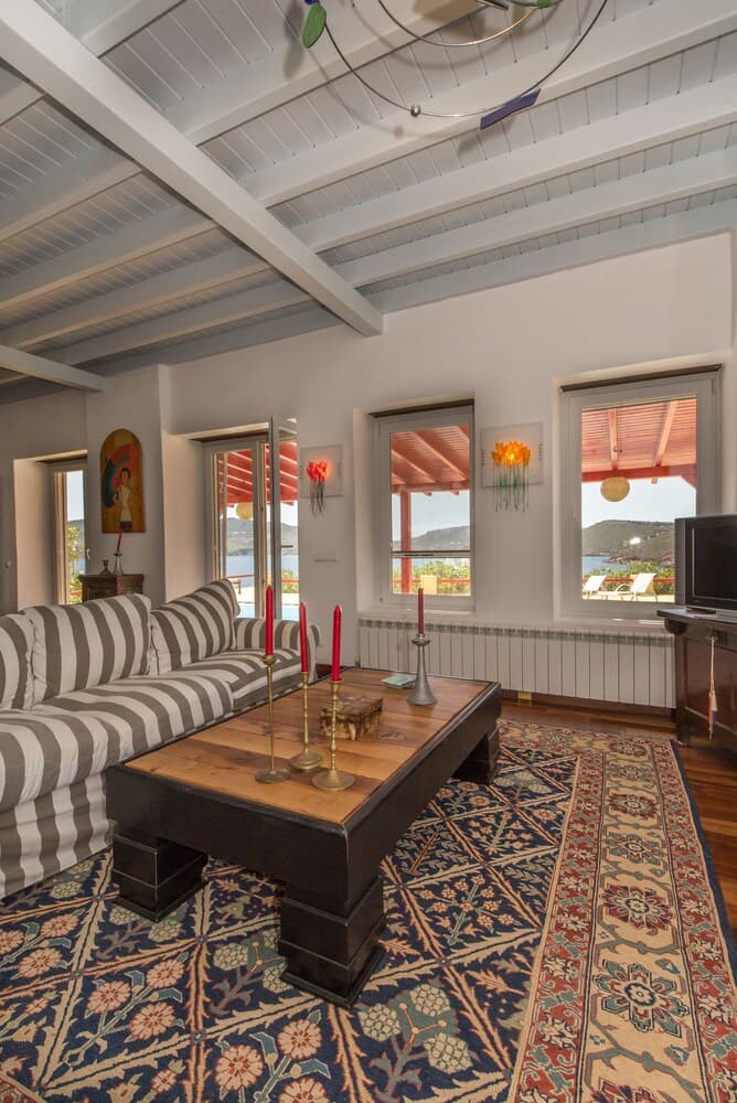 A cozy living room with a striped sofa, wooden coffee table adorned with red candles, and a vibrant rug, framed by large windows offering scenic views of the sea, ideal for travelers seeking a serene retreat in a charming coastal villa.