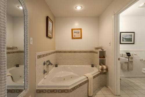 A cozy hotel bathroom features a corner bathtub with tiled surround, framed artwork on the walls, and a mirror reflecting the serene ambiance, ideal for travelers seeking relaxation.