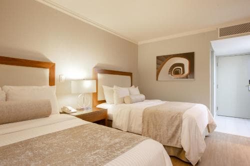 A cozy hotel room features two neatly made beds with beige linens, a modern bedside lamp, and abstract wall art, creating a serene atmosphere ideal for travelers seeking relaxation.