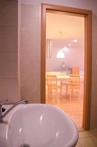 A cozy apartment bathroom features a white sink in the foreground, leading to a warmly lit dining area with wooden chairs and a table, ideal for travelers seeking comfortable accommodations.